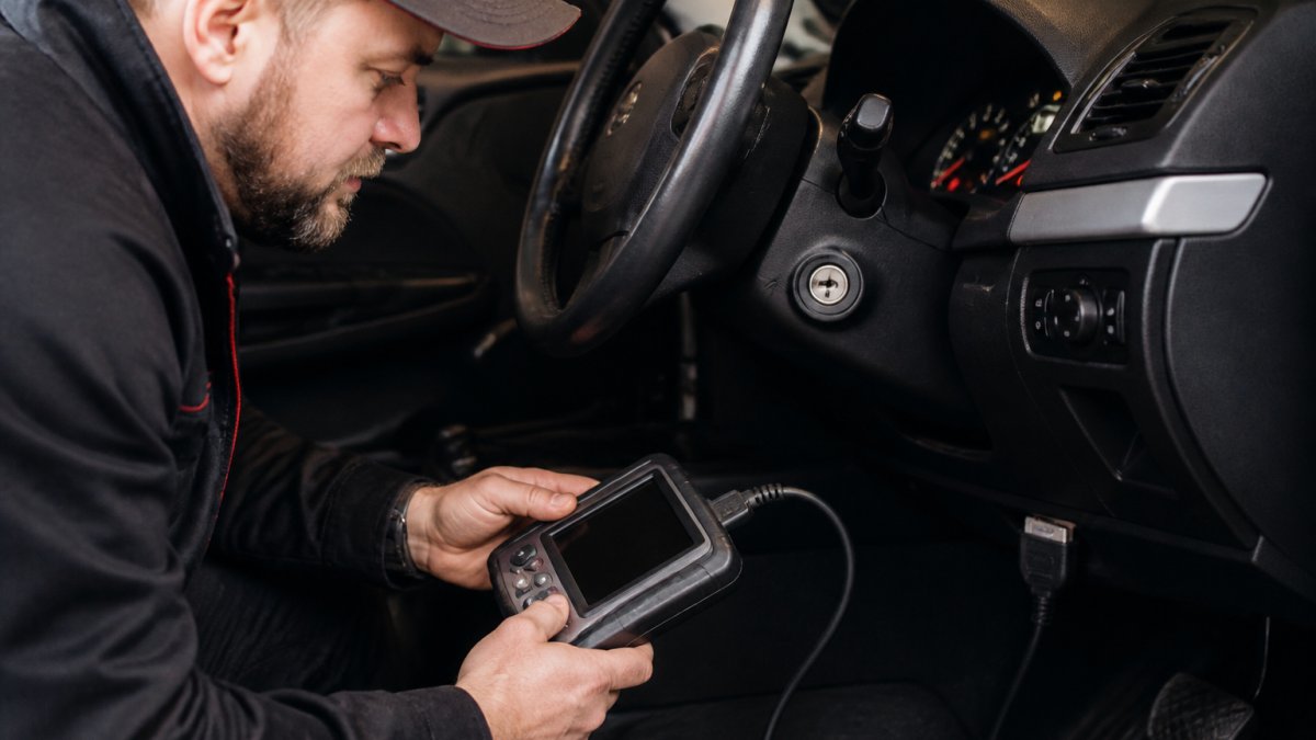 An automotive locksmith working on a car's dashboard area at night, holding a diagnostic scan tool connected to the OBD port, with the vehicle's security light glowing on the instrument cluster