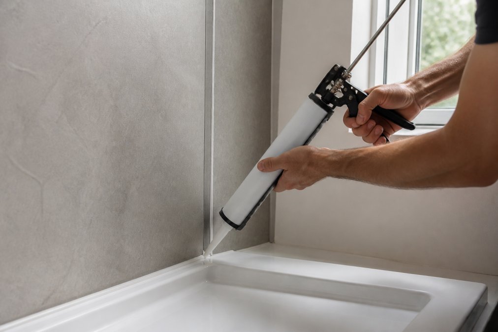 Professional hand applying silicone sealant along a panel joint using a caulking gun, showing a smooth, continuous bead application in a modern bathroom setting with natural window lighting