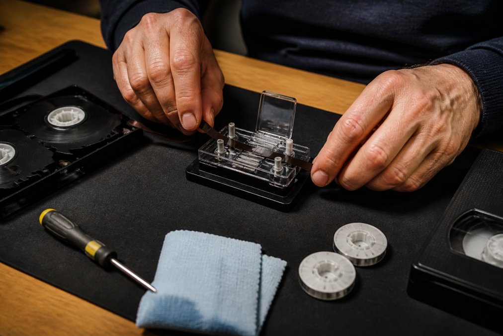 Close-up of hands carefully using a splicing block to repair a broken VHS tape on a clean, well-lit workbench, with a screwdriver and cleaning cloth nearby