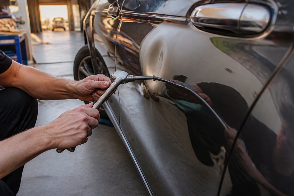 Close-up of a certified technician's hands using a PDR tool to gently massage a large dent on a car door, the vehicle's paint visibly undamaged and glossy in the late afternoon sunlight