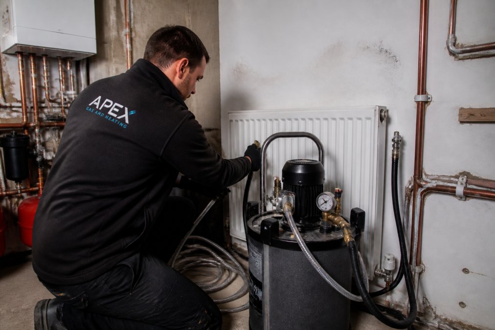 Professional heating engineer operating a power flushing machine connected to a radiator system with visible hoses and gauges, demonstrating the equipment in action in a residential boiler room