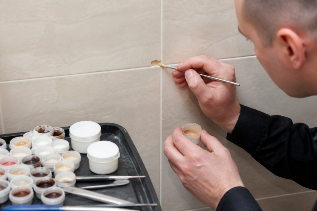 Close-up of a technician's hands applying colour-matched filler to a chipped porcelain wall tile, with a palette of pigments and precision tools on a nearby tray, bright task lighting