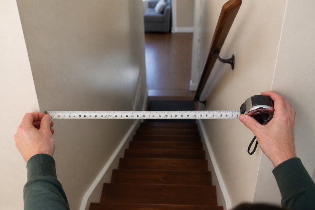 Close-up of a person's hands using a steel tape measure to measure the width of a narrow residential staircase, with the tape clearly showing measurement markings at the staircase opening between two walls