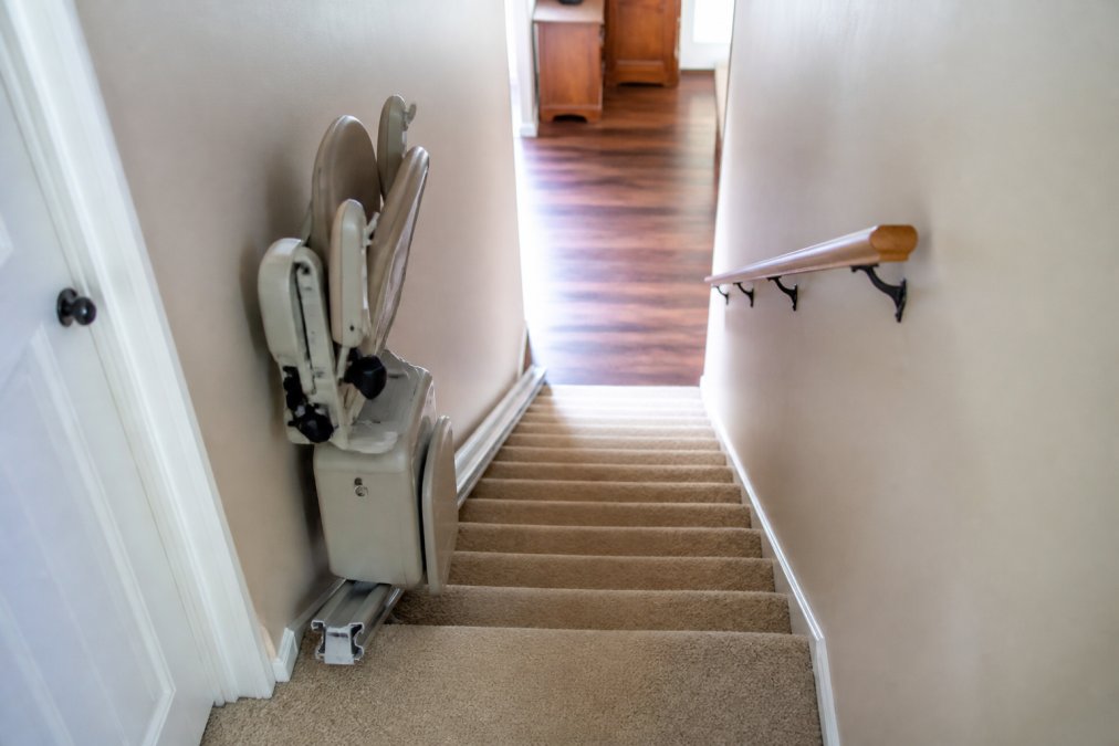 An installed residential stairlift on a narrow staircase with the seat folded against the wall, showing clear 24-inch clearance on the opposite side of the stairs and the track mounted flush against the wall in a tight hallway