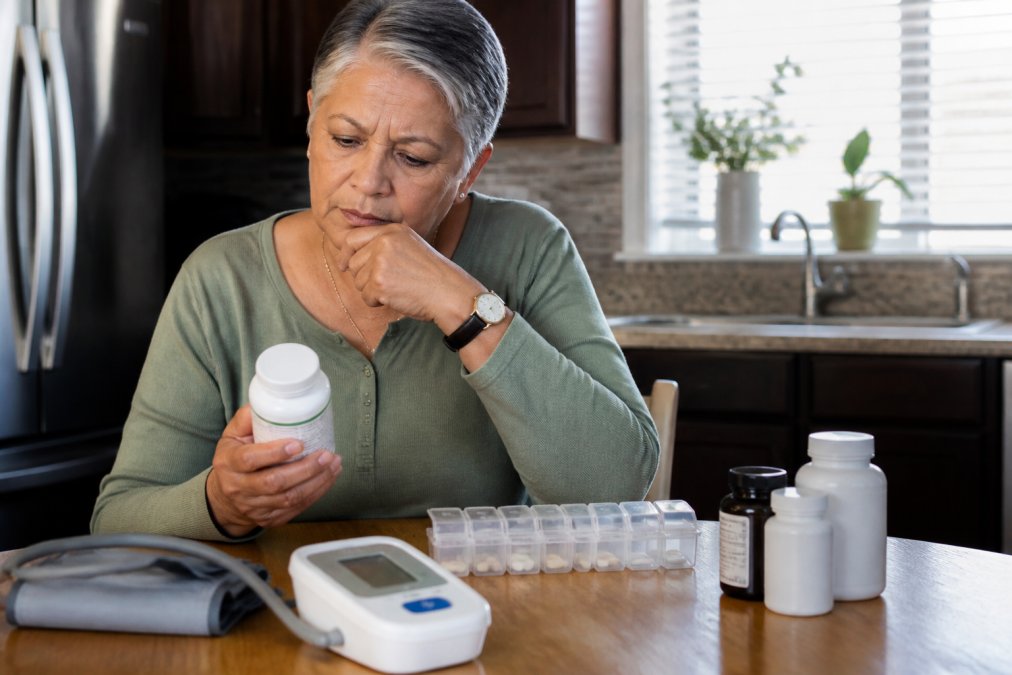 A person sitting at a kitchen table with a pill organizer, a blood pressure monitor, and several supplement bottles, looking thoughtfully at the labels