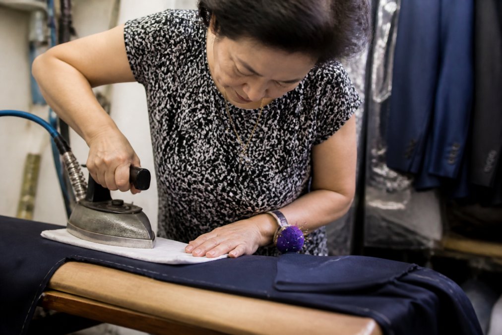 A professional tailor using a pressing iron and press cloth on a suit jacket seam on a wooden pressing board with proper lighting, demonstrating meticulous technique