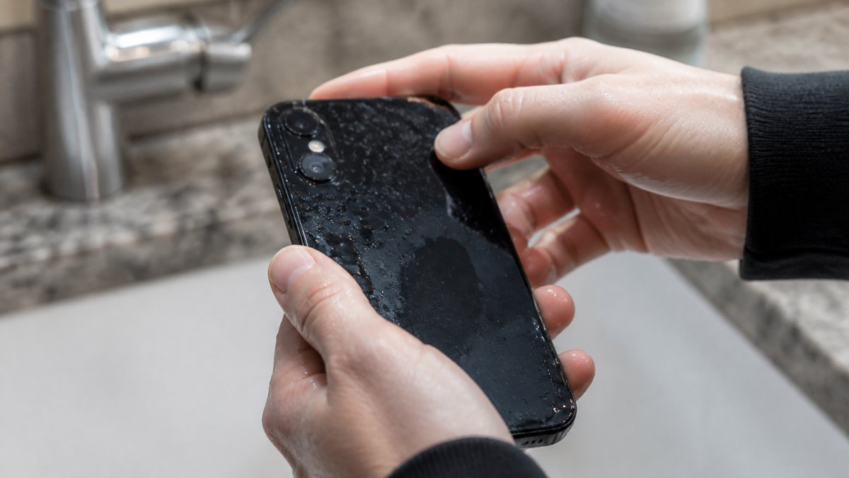 Close-up of hands holding a wet smartphone over a sink with water droplets visible on the screen and back of the device, fingers positioned to power it off