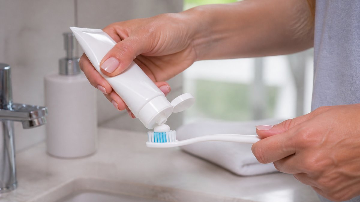 Close-up of a person applying desensitising toothpaste to their toothbrush, with a toothbrush and toothpaste tube visible on a bathroom counter