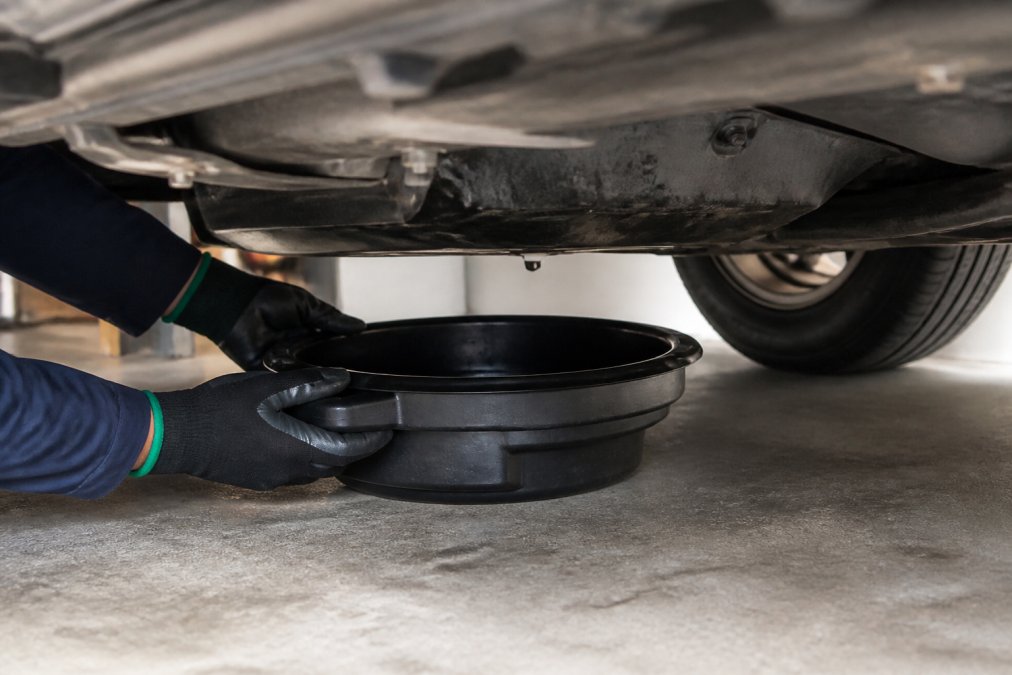 Hands in work gloves positioning a drain pan under a vehicle's drain plug, showing proper technique and safe positioning beneath the engine