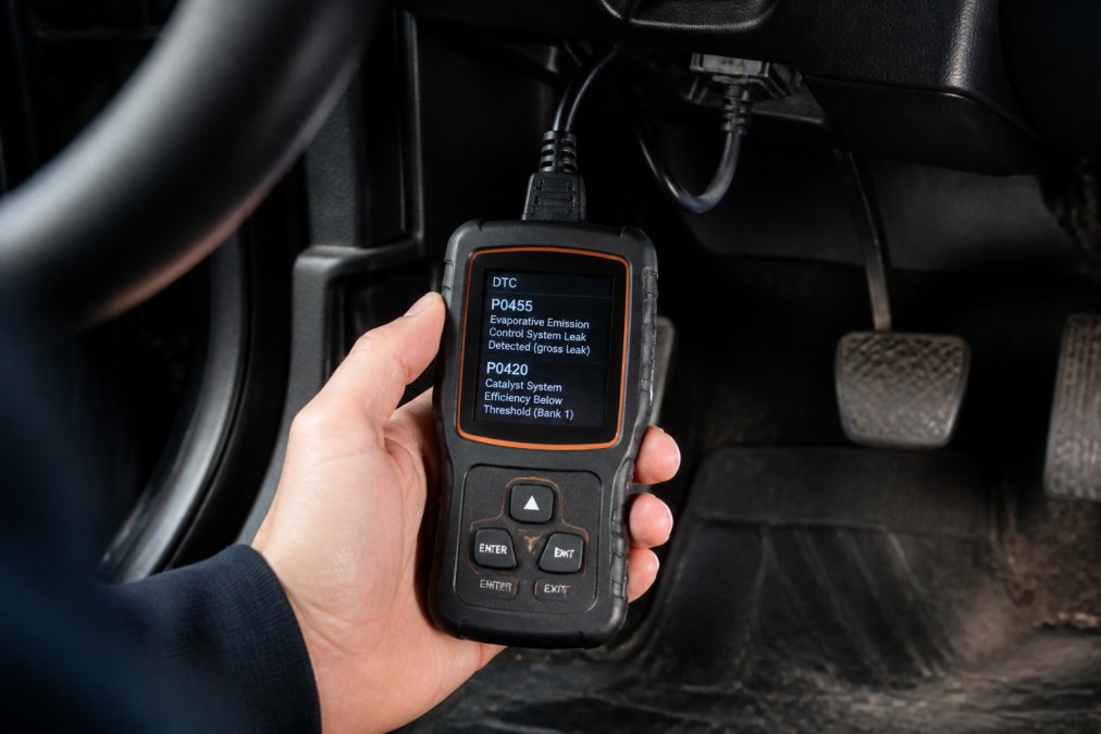 Close-up of hands holding an OBD-II scanner device plugged into a vehicle's diagnostic port beneath the dashboard, with the scanner screen displaying fault codes in bright digital text