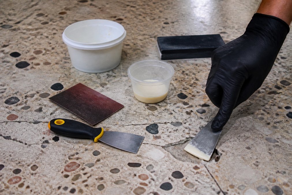 Close-up of a terrazzo floor with a visible chip, surrounded by repair tools including a putty knife, epoxy, and sandpaper, with a gloved hand applying epoxy to the chip