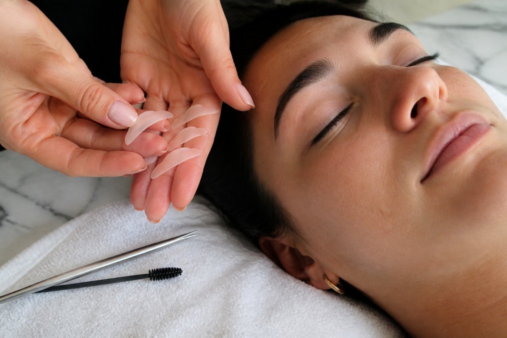 Close-up of a lash technician's hands holding small silicone lifting shields of different sizes next to a model's closed eye, with a clean white towel and lash tools on a marble counter
