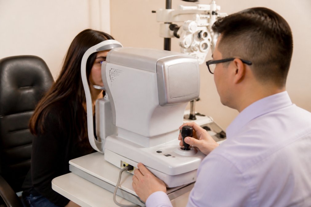 Optometrist using advanced measurement equipment to examine a patient's eye during contact lens fitting, with patient seated in examination chair under warm clinical lighting
