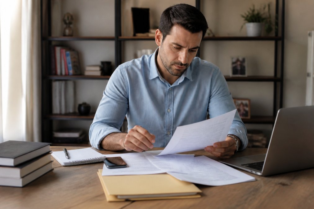 A person sitting at a desk with legal papers, a phone, and a laptop, looking focused and determined while reviewing court documents in a home office