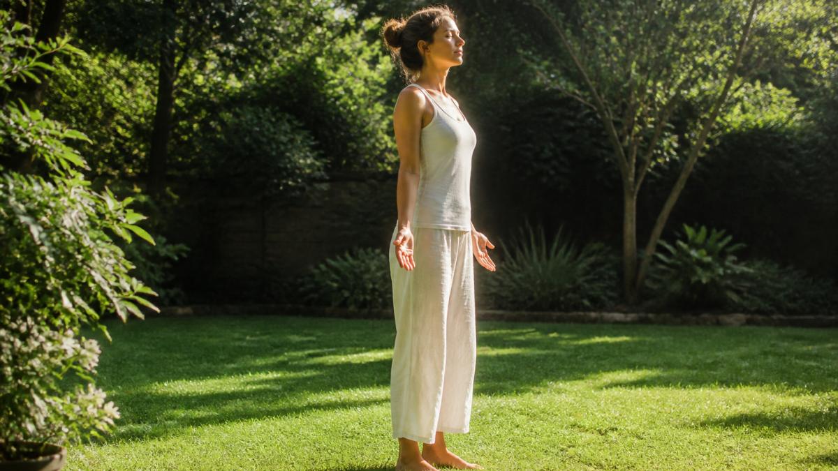 A person standing barefoot on grass in a sunlit garden, eyes closed, hands relaxed at their sides, warm morning light filtering through trees