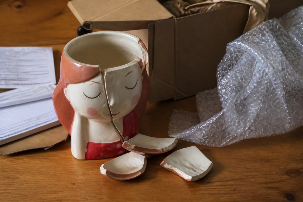 Close-up of damaged ceramic pot with broken pieces and cracks visible, photographed next to original packaging materials, bubble wrap, and order documentation on a wooden surface