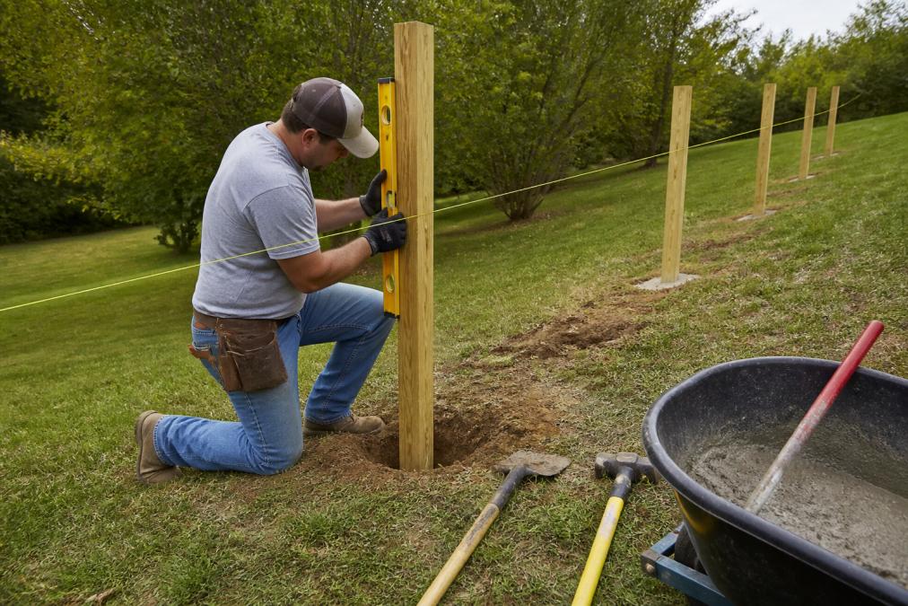 A fence installer in work clothes using a level and string line to set a wooden post on a sloped backyard, with concrete mix and digging tools nearby