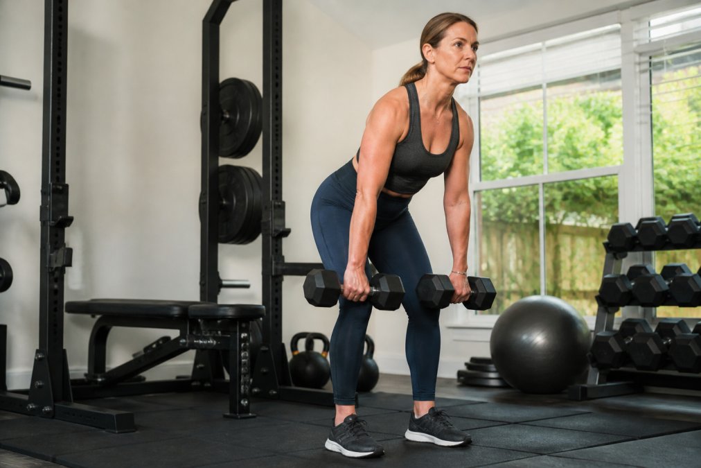 A woman in her mid-40s performing a dumbbell deadlift in a bright, tidy home gym, focused expression, natural light coming through a window behind her