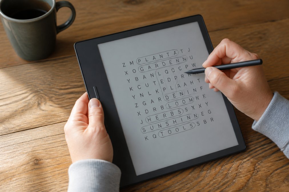 Close-up of hands using a stylus to write answers on a Kindle Scribe displaying a word search puzzle grid, wooden desk surface visible in the background, soft natural light