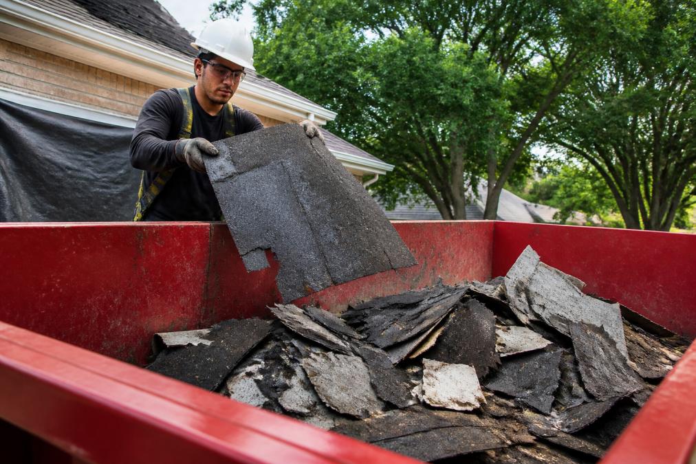 Worker in safety gear loading asphalt debris into a residential roofing shingles roll-off dumpster