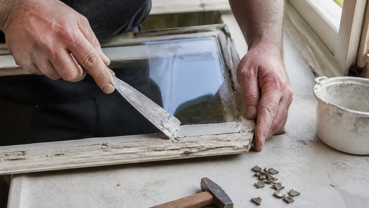 Close-up of a glazier's hands using a putty knife to press new putty around a freshly fitted glass pane in a timber window frame, with a small hammer and glazing points resting on a windowsill nearby