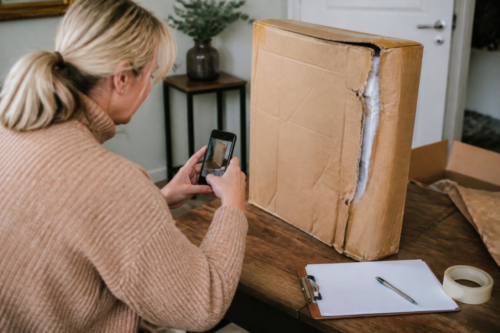 Person inspecting a damaged painting box with visible dents and tears, taking photos with a smartphone to document damage for a return claim on a home desk