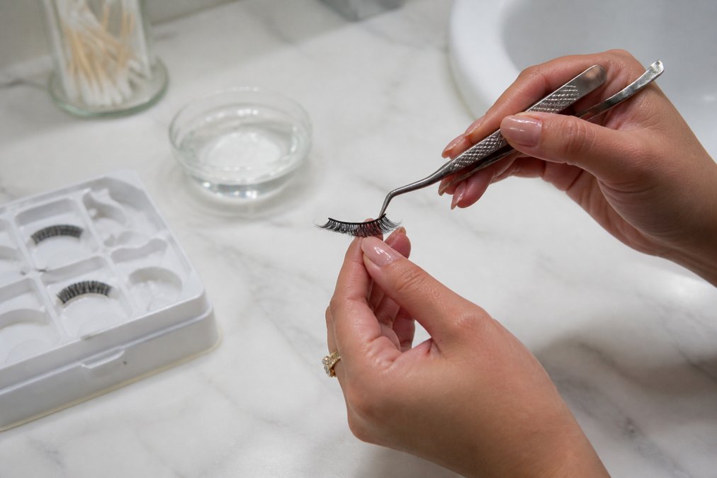 Using tweezers to gently remove adhesive to safely reuse press-on eyelashes over a bathroom counter.
