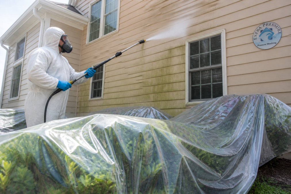 Technician in protective gear soft washing a house with shrubs covered in plastic to safely use bleach.