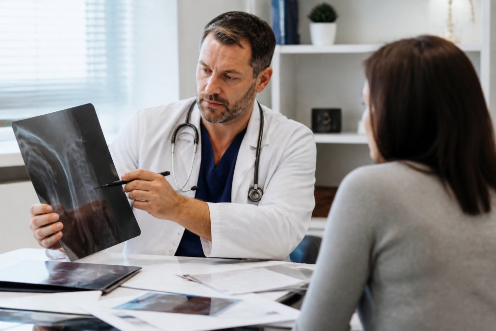 Doctor reviewing X-rays and medical records with patient in clinical office setting, papers spread on desk with imaging results visible