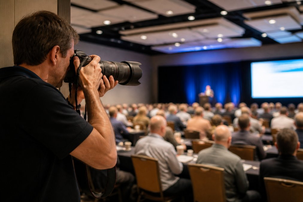 A photographer holding a DSLR camera with a long telephoto lens, positioned at the edge of a bright conference hall, attendees blurred in the background