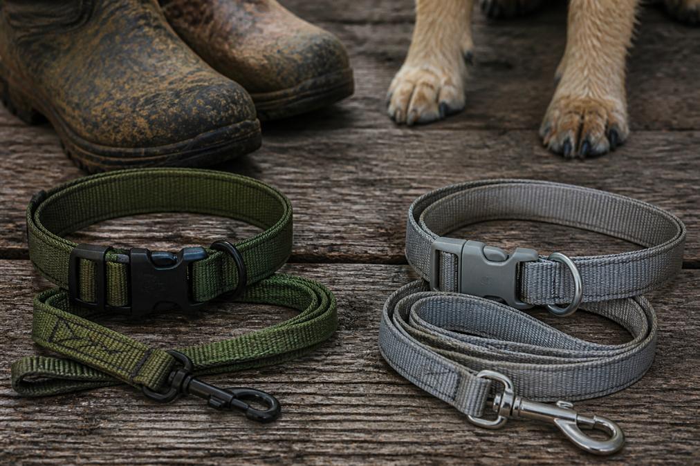 Close-up of a rugged nylon dog collar and leash resting on a weathered wooden surface next to a pair of muddy hunting boots, with a dog's paw visible in the background