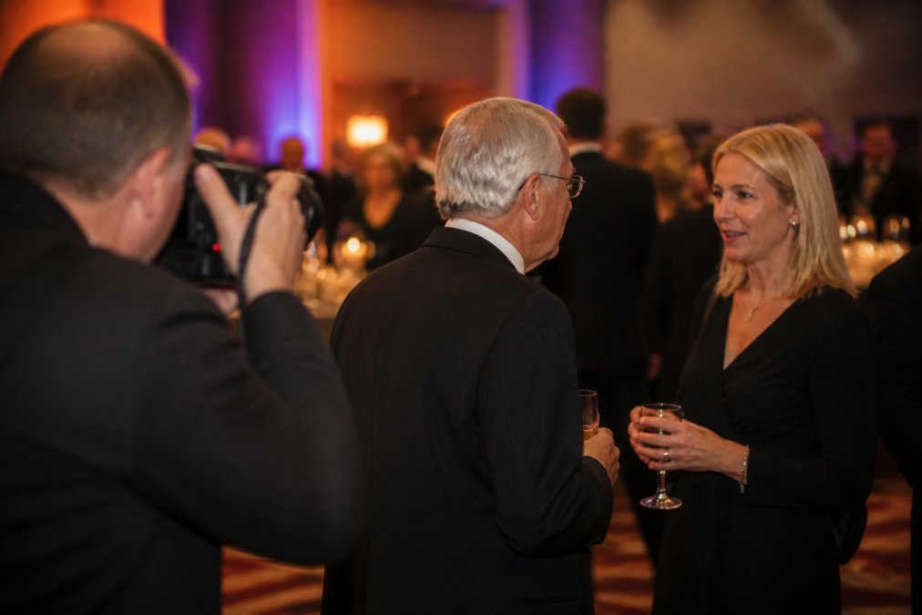Professional photographer discreetly capturing a genuine moment between a nonprofit executive and a major donor in conversation at a gala event, with elegant lighting and other attendees softly blurred in the background
