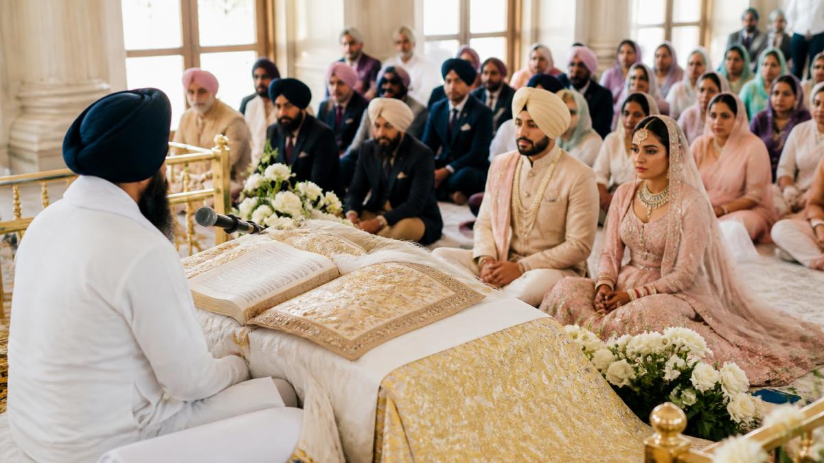 A Sikh bride and groom seated respectfully before the Guru Granth Sahib in a Gurdwara, the Granthi reading from the holy book, family gathered around, soft natural light filtering through windows