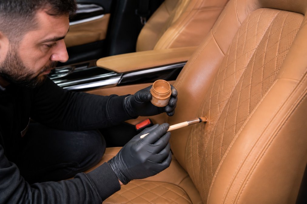 Professional craftsperson's hands carefully applying premium leather treatment to a luxury car seat using precision tools and colour-matched restoration materials in a well-lit workshop