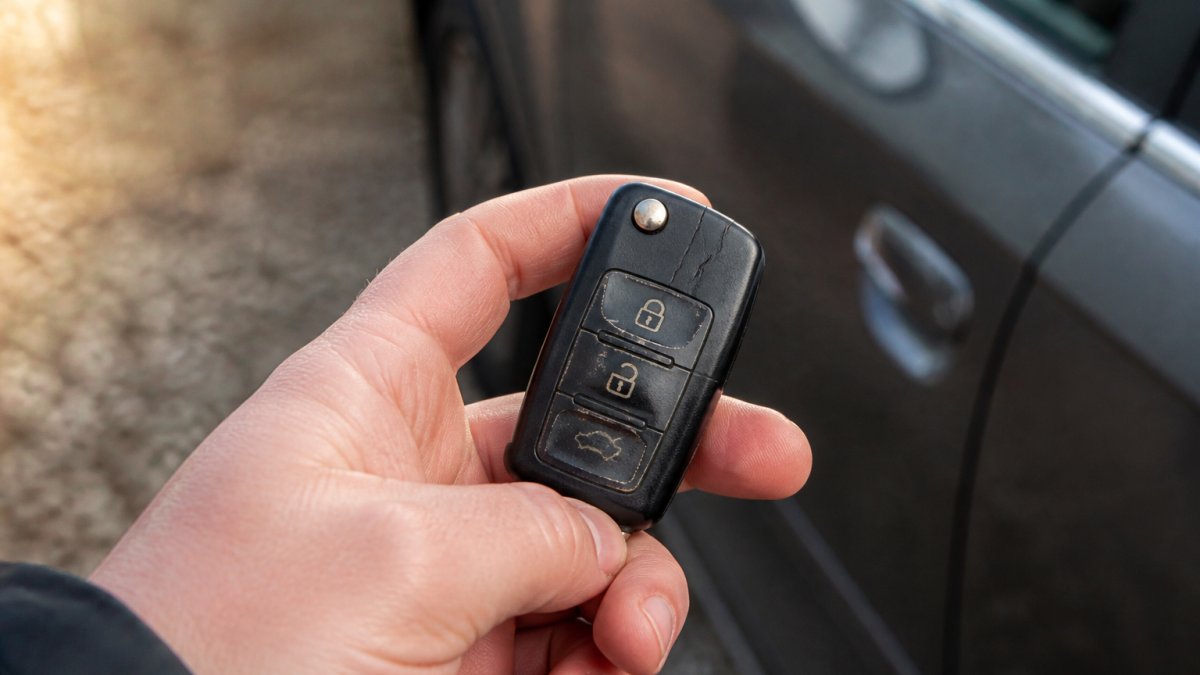 Close-up of a car key fob held in a person's hand, worn buttons and a hairline crack visible on the casing, driveway gravel and a car door in soft background light
