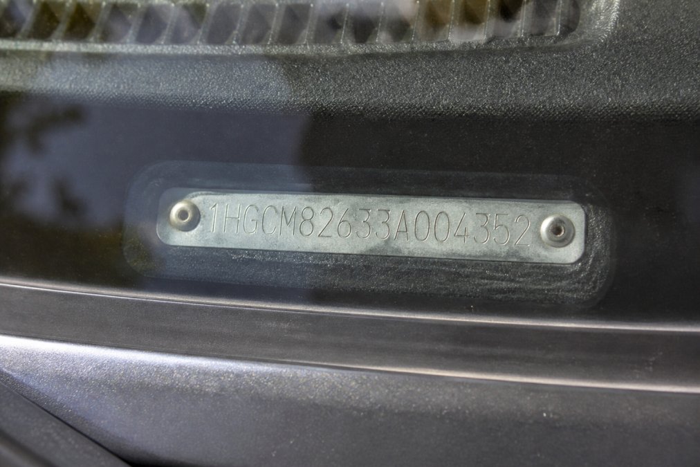 Close-up of a VIN plate mounted on a car's dashboard visible through the windshield, showing the 17-character identification number clearly etched into the metal plate under bright daylight