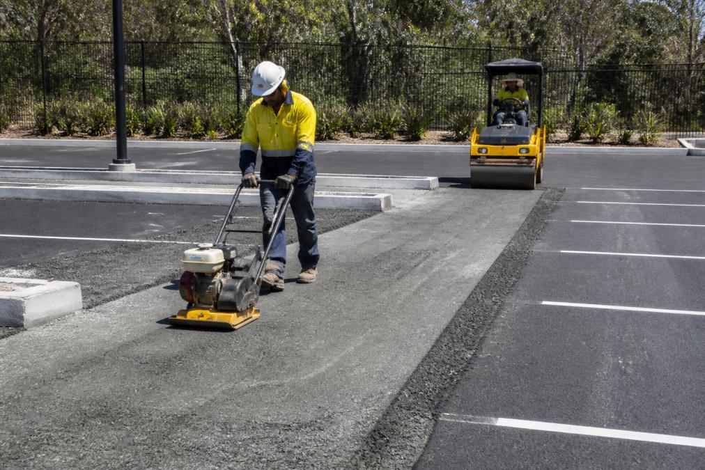 Paving crew using a plate compactor to install a base for commercial car park paving with a roller nearby.