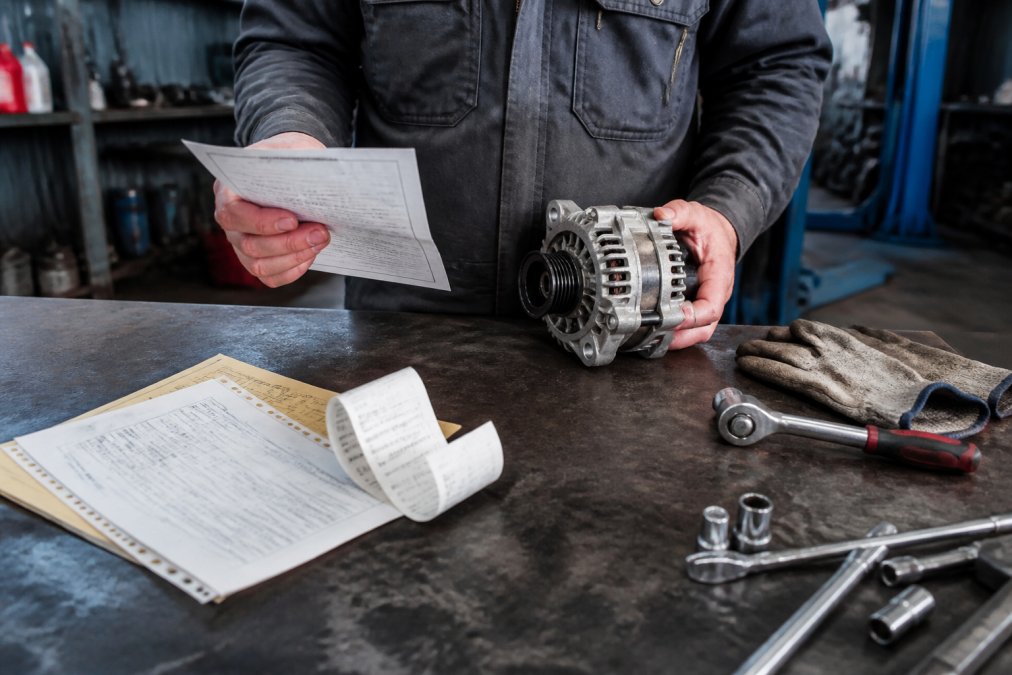 Person at a workbench holding warranty documentation and a replaced car part, with service records and receipt visible on the surface