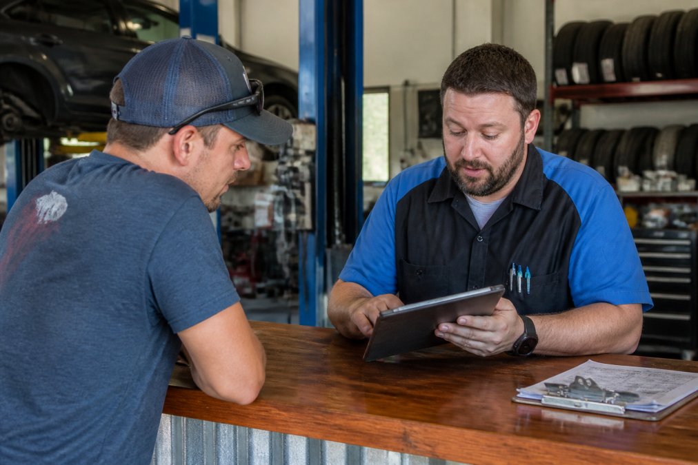 Customer sitting at service counter reviewing repair invoice with technician on tablet, discussing payment options in modern automotive shop