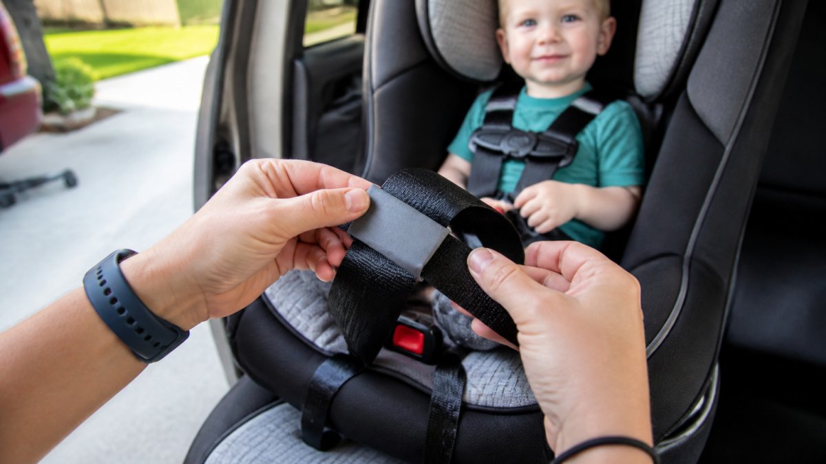 Parent threading a car seat harness through a silicone strap protector in a sunny driveway