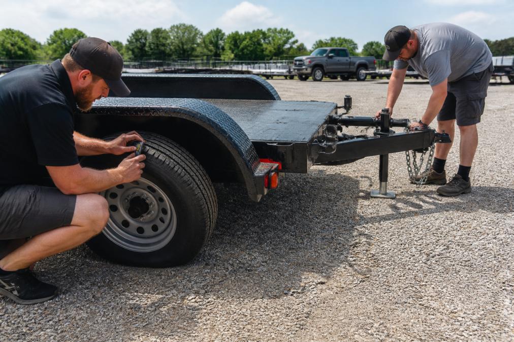 A person kneeling beside a rental car trailer, checking the tire tread depth with a gauge while another person inspects the coupler latch and safety chains in a bright outdoor lot