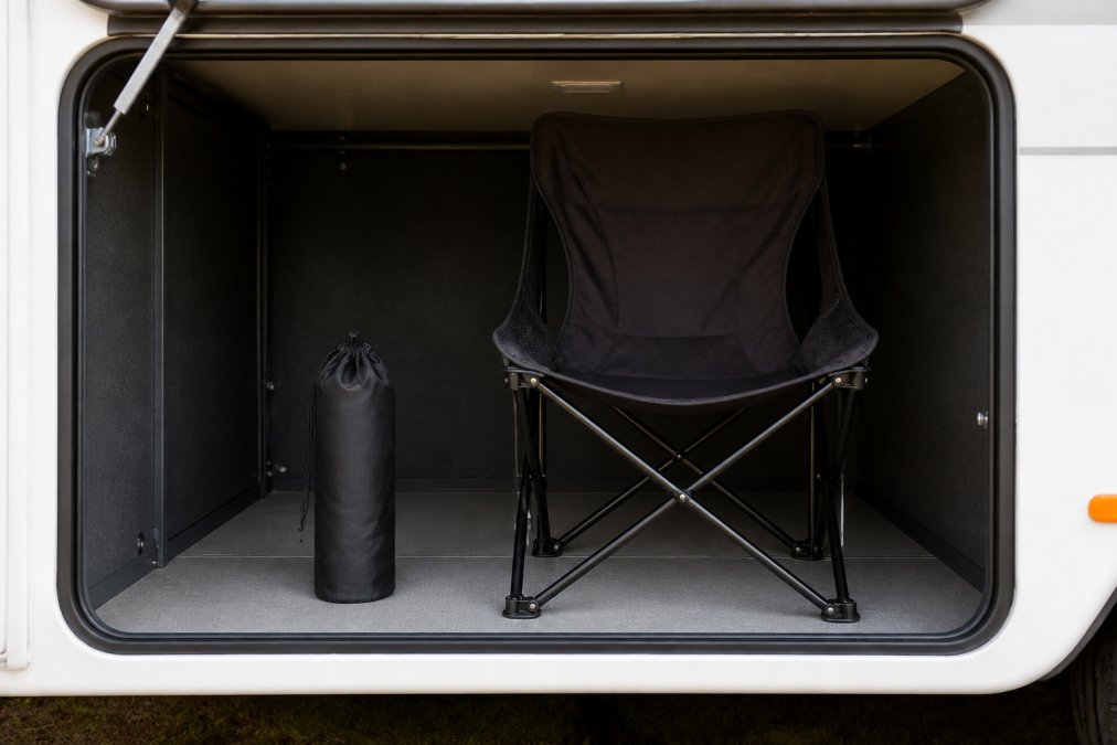 A compact black camping chair folded to drink-bottle size sitting next to a full-size unfolded chair inside an open caravan storage locker, showing the space saved