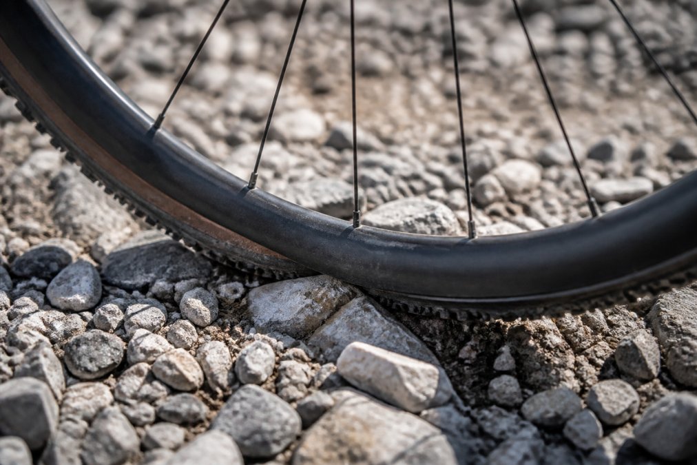 Close-up of a gravel bike wheel being tested on rough rocky terrain, showing the rim contact with jagged stones and the spoke tension under lateral stress in bright daylight