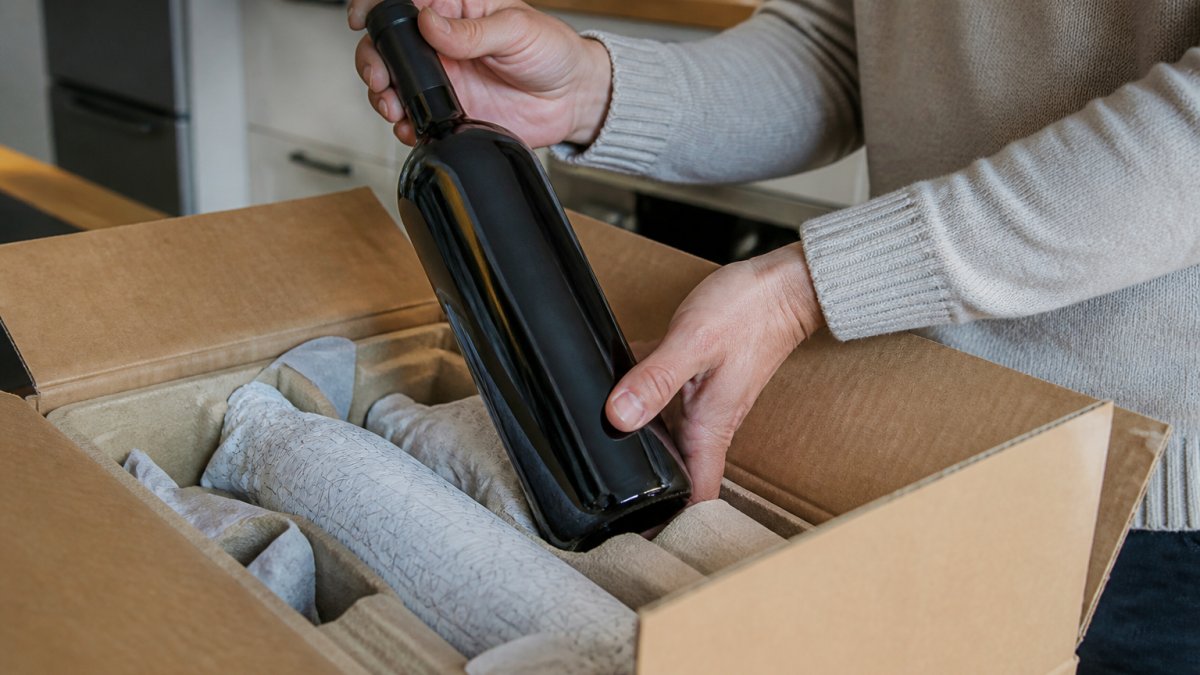 Close-up of hands lifting a wine bottle from a cardboard delivery box filled with moulded pulp inserts and protective wrapping, on a kitchen table