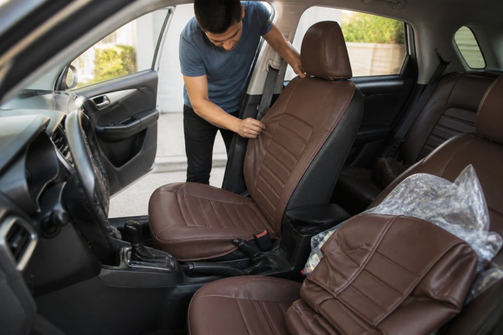 A person installing a brown leather-look seat cover on the front seat of a compact car parked in a sunny driveway, with a second seat cover and packaging visible on the passenger side