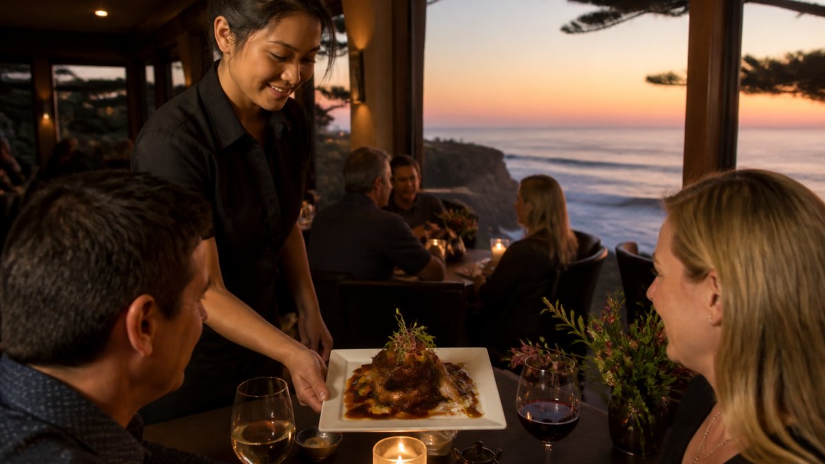 Server presenting an elegantly plated Asian fusion dish at a candlelit table overlooking Carmel's coastal landscape, with warm ambient lighting and guests dining in the background