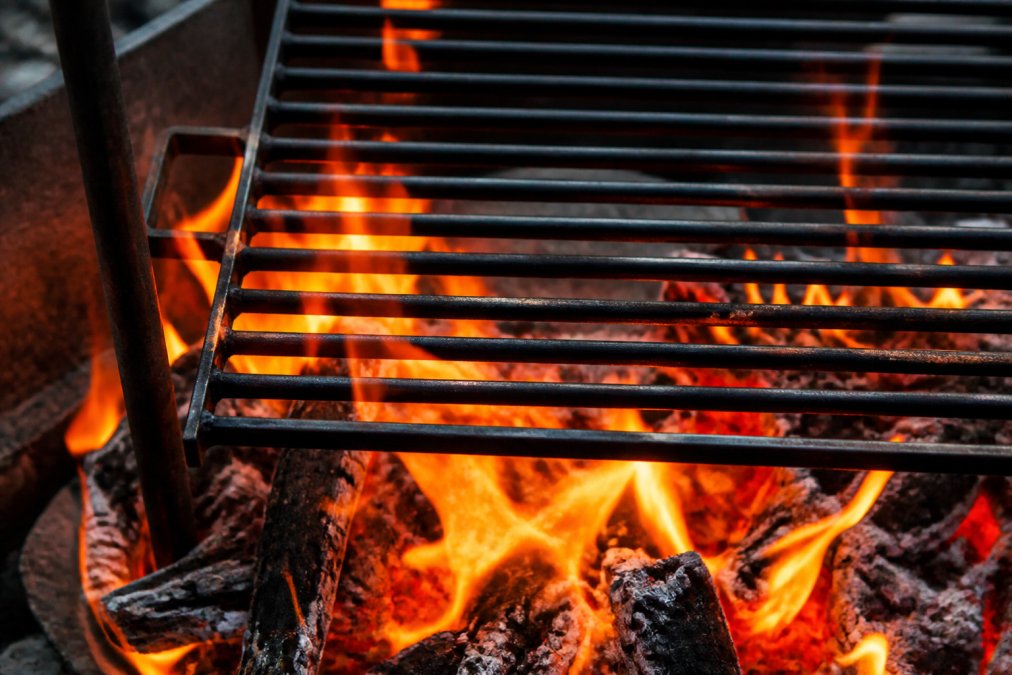Close-up of a heavy carbon steel grill grate over an active campfire with flames licking around the metal, showing direct heat contact and the characteristic orange glow of coals beneath