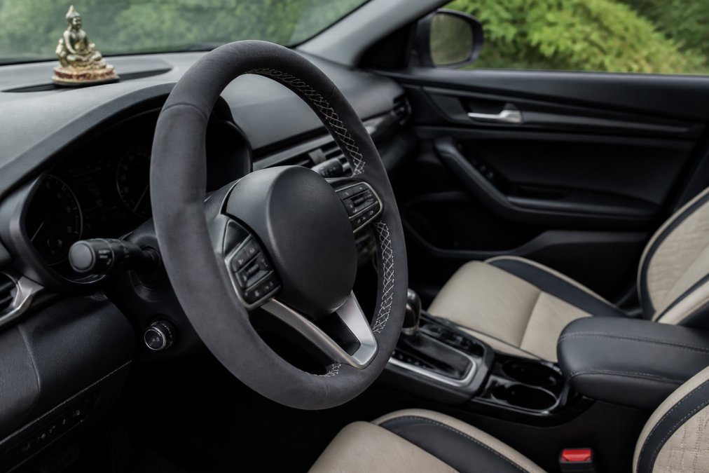 Close-up of car interior showing a hand-stitched suede leather steering wheel cover installed on a steering wheel, with custom seat covers visible in the background, and a decorative car ornament on the dashboard, all in a modern vehicle cabin with natural lighting