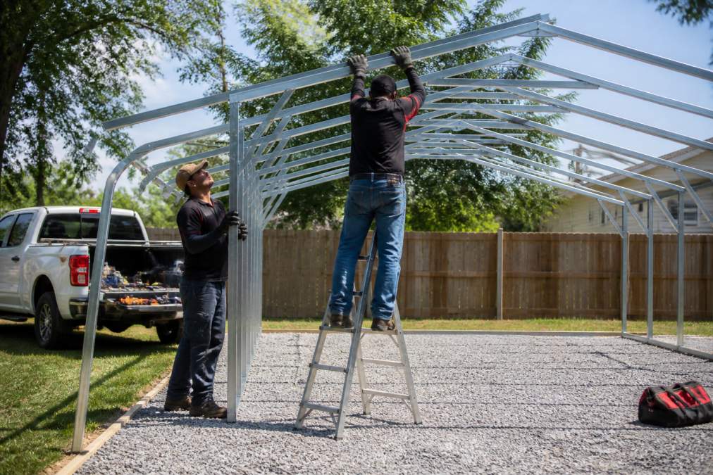 A professional installation crew assembling a galvanized steel carport frame on a prepared gravel pad, with a truck and tools visible in a residential backyard setting
