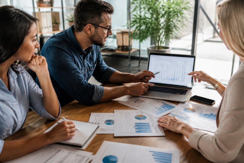 Marketing team reviewing case study results and performance metrics on laptop screen, with printed analytics reports and performance data visible on desk in modern office
